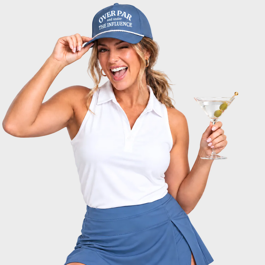 A woman in a white sleeveless polo shirt and blue skirt wears a blue cap that reads 'OVER PAR THE INFLUENCE' and holds a martini glass against a light gray studio background.